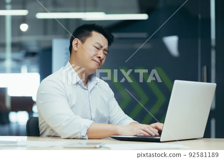 A young man, an Asian businessman at work in the office, sits at the table, holds his neck, feels pain, closes his eyes, A young man, an Asian businessman at work in the office, sits at the table, holds his neck, feels pain, closes his eyes, 92581189