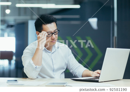 Serious and focused young Asian businessman in glasses, freelancer, entrepreneur. Sitting at a table in a modern office, working with a laptop and documents. Serious and focused young Asian businessman in glasses, freelancer, entrepreneur. Sitting at a table in a modern office, working with a laptop and documents. 92581190