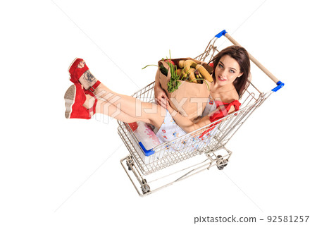 High angle view of girl smiling at camera while holding grocery bags and sitting in shopping trolley isolated on white High angle view of girl smiling at camera while holding grocery bags and sitting in shopping trolley isolated on white 92581257
