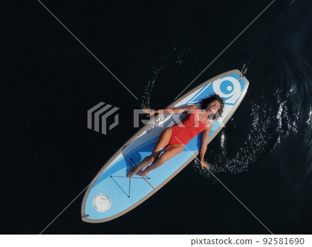 Aerial drone view on well looking middle aged woman with black hair in red swimsuit, swimming on sup in calm azure sea. Summer holiday vacation and travel concept. Aerial drone view on well looking middle aged woman with black hair in red swimsuit, swimming on sup in calm azure sea. Summer holiday vacation and travel concept. 92581690