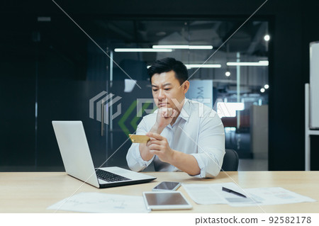 Worried and pensive young man Asian businessman is holding a credit card in his hands, looking at it. Working on a laptop. Sitting in the office at the desk 92582178
