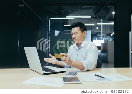 Worried young Asian businessman looking surprised at laptop and credit card in hand. Sitting in the office at the desk Worried young Asian businessman looking surprised at laptop and credit card in hand. Sitting in the office at the desk 92582182