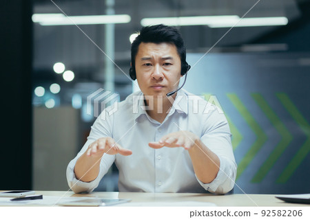 Business coach. A young handsome Asian man in headphones with a microphone conducts business training on camera, explains, tells, smiles. Sitting behind a desk in a modern office. 92582206