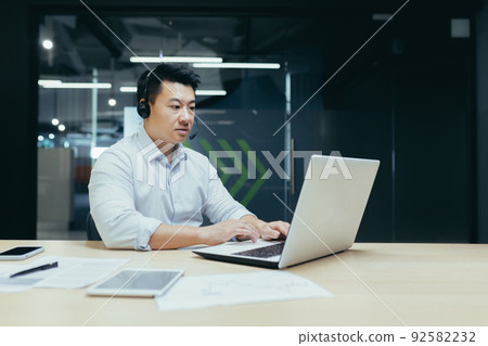 Young handsome Asian businessman working on laptop in headphones with microphone. Sitting at a desk in a modern office. Serious, focused. 92582232