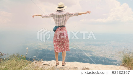 Woman traveler flaps arms like wings on mountain top. Young woman in hat opens arms wide waving like bird's wings over Tira city, Albania. Red dress happy girl blowing wind. Dream, travel concept 92582367