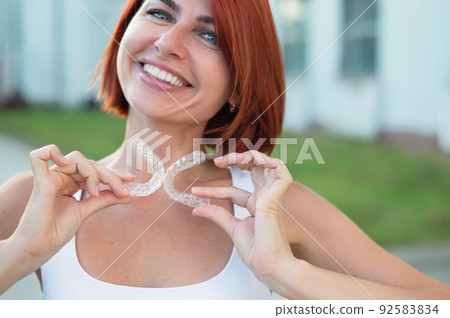 Red-haired Caucasian woman holding transparent mouthguards for bite correction outdoors. A girl with a beautiful snow-white smile uses silicone braces 92583834