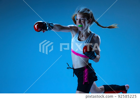Junior league. Young girl MMA fighter practicing punch isolated on blue background in neon light. Concept of sport, competition, action, achievements Junior league. Young girl MMA fighter practicing punch isolated on blue background in neon light. Concept of sport, competition, action, achievements 92585225