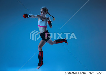 Dynamic portrait of young girl, junior MMA fighter in action, motion isolated on blue background in neon light. Concept of kids sport Dynamic portrait of young girl, junior MMA fighter in action, motion isolated on blue background in neon light. Concept of kids sport 92585226
