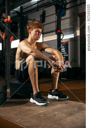 Portrait of young sportive man sitting on weights and resting isolated over gym background. Training 92589205