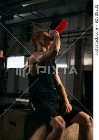 Portrait of young sportive man in black uniform sitting on box, drinking water, having break isolated over gm background Portrait of young sportive man in black uniform sitting on box, drinking water, having break isolated over gm background 92589232