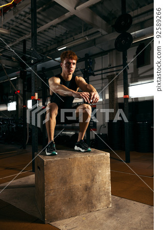 Portrait of sportive, muscular red-haired young man jumping on box, doing exercises isolated over gym background Portrait of sportive, muscular red-haired young man jumping on box, doing exercises isolated over gym background 92589266