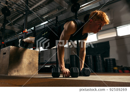 Portrait of young muscular red-headed man doing push up exercises with dumbbells isolated on indoor gym background Portrait of young muscular red-headed man doing push up exercises with dumbbells isolated on indoor gym background 92589269