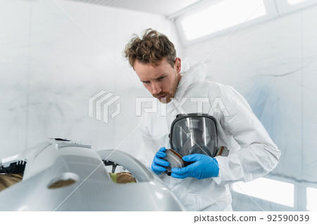 Car paint service worker in a protective suit and a mask in his hands checks the quality of the painting and varnishing works work performed 92590039