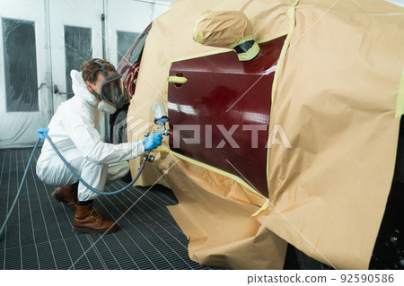 Auto painter in a protective suit and mask sprays a degreaser or other cleaner on a car door using a spray gun. Preparation of vehicle body elements for local repair 92590586