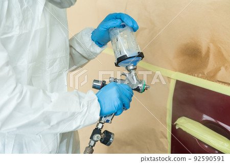 Close up view of auto painter in a protective suit and mask sprays a degreaser or other cleaner on a car door using a spray gun. Preparation of vehicle body elements for local repair 92590591