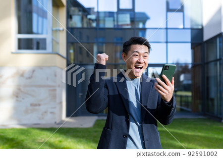 Exited Asian Businessman Celebrating Achievement looking at Smartphone outdoor. Successful business man celebrating win with mobile phone outside. Happy Good News. Successful Deal, Startup Bet 92591002