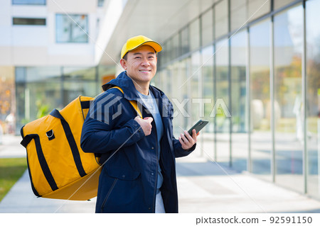 Happy male courier walking food supplier, looking forward to new order received in phone app, asian courier Happy male courier walking food supplier, looking forward to new order received in phone app, asian courier 92591150