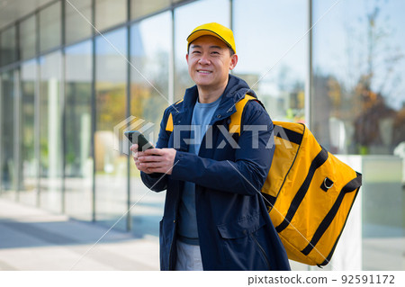 Happy male courier walking food supplier, looking forward to new order received in phone app, asian courier 92591172
