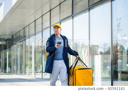 asian male food courier using mobile using mobile phone app on smartphone while standing city street with insulated with thermal backpack delivering food. looking customer address. purchase online map 92591181