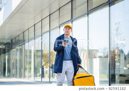 asian male food courier using mobile using mobile phone app on smartphone while standing city street with insulated with thermal backpack delivering food. looking customer address. purchase online map 92591182