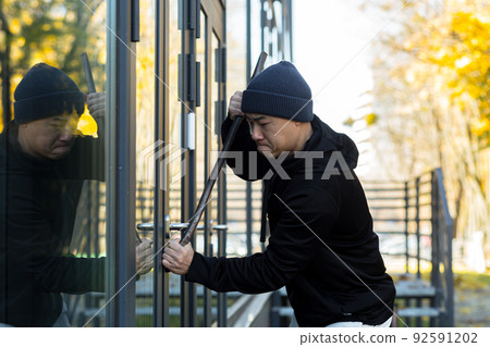 Asian man robber tries to break the door lock with a large metal crowbar tool 92591202