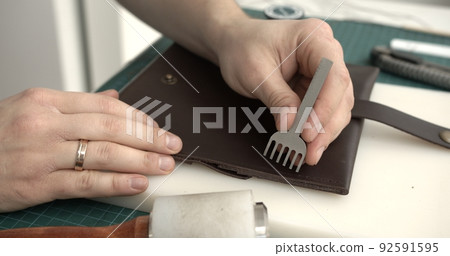 Craftsman making holes in leather with puncher tool. Working process with a brown natural leather. Craftsman making holes in leather with puncher tool. Working process with a brown natural leather. 92591595