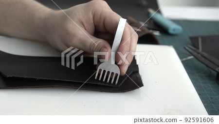 Close up men's hands make a leather product. Punching holes in the leather with a hole punch with a hammer. Tools for leather craft. 92591606