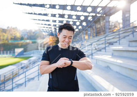 Sport asian man looking smart watch at modern track. Man runner using smartwatch bracelet. Portrait fitness man checking result. Male in sportswear jogging in the street urban background city stadium 92592059