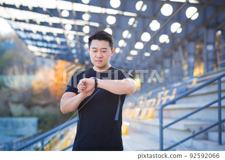 Sport asian man looking smart watch at modern track. Man runner using smartwatch bracelet. Portrait fitness man checking result. Male in sportswear jogging in the street urban background city stadium 92592066