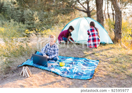 Lets go camping. Family camping. Reach destination place. Two girls pitch tent and one boy watching show on the laptop, sitting on the plaid. Wanderlust discovery. Hiking outdoor adventure. Lets go camping. Family camping. Reach destination place. Two girls pitch tent and one boy watching show on the laptop, sitting on the plaid. Wanderlust discovery. Hiking outdoor adventure. 92595977