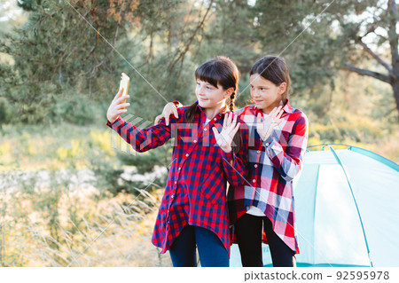 Lets go camping. Family camping. Reach destination place. Cheerful teenage girls talking on a video call, looking at the phone screen in the middle of a pine forest near tent. Wanderlust discovery. Lets go camping. Family camping. Reach destination place. Cheerful teenage girls talking on a video call, looking at the phone screen in the middle of a pine forest near tent. Wanderlust discovery. 92595978
