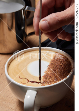 Top view shot of professional barista pouring milk from jar in to a cup of coffee, coffee being prepared by a barista. Focus on hands holding cup of coffee. 92597257