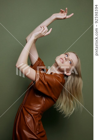 Curving in dance blonde young female model in brown trendy dress holding hands up dreaming looks up aside posing isolated on over olive green studio wall background. Seasonal sale banner 92598394