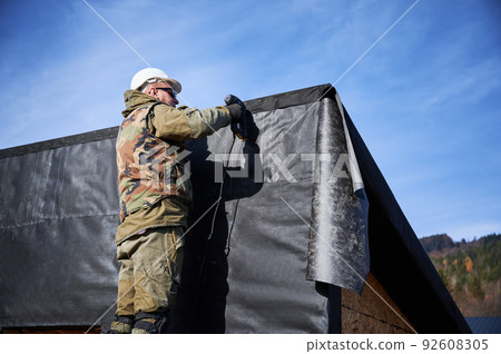 Male builder installing waterproof membrane on the wall of future cottage. Man worker building wooden frame house with blue sky on background. Carpentry and construction concept. 92608305