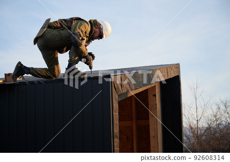 Male builder installing black corrugated iron sheet used as facade of future cottage. Man worker building wooden frame house. Carpentry and construction concept. 92608314