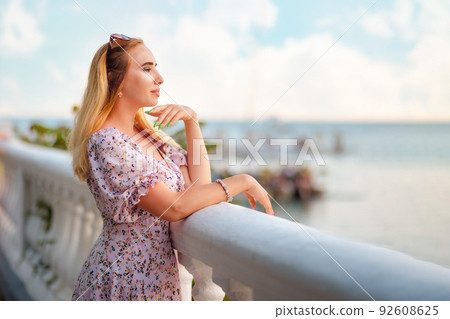 Portrait of a beautiful blonde woman posing at the balustrade and looking into the ocean. Copy space. The concept of summer vacation, freedom and psychology Portrait of a beautiful blonde woman posing at the balustrade and looking into the ocean. Copy space. The concept of summer vacation, freedom and psychology 92608625