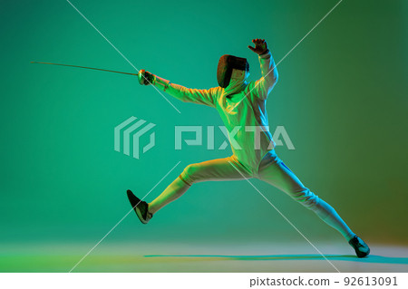 Studio shot of young man, fencer with smallsword practicing fencing isolated on green background in neon light. Sport, energy, skills, achievements 92613091