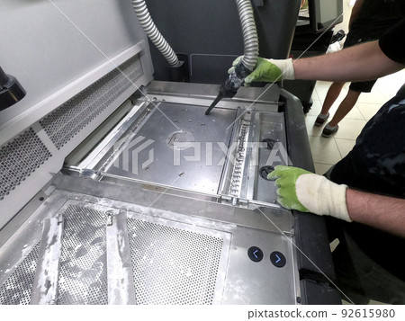 A male worker cleans the surface of an industrial 3D printer from white powder with a vacuum cleaner close-up. Multi Jet Fusion MJF 3D printing. The working process A male worker cleans the surface of an industrial 3D printer from white powder with a vacuum cleaner close-up. Multi Jet Fusion MJF 3D printing. The working process 92615980