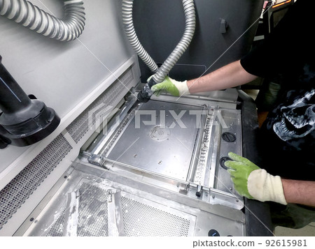 A male worker cleans the surface of an industrial 3D printer from white powder with a vacuum cleaner close-up. Multi Jet Fusion MJF 3D printing. The working process A male worker cleans the surface of an industrial 3D printer from white powder with a vacuum cleaner close-up. Multi Jet Fusion MJF 3D printing. The working process 92615981