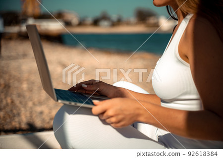 young beautiful woman with dark hair in white overalls sits on the sand near the sea and uses a laptop young beautiful woman with dark hair in white overalls sits on the sand near the sea and uses a laptop 92618384