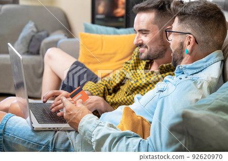 Happy young gay couple using laptop computer while sitting on a couch at home, shopping online with 92620970