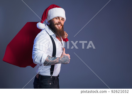 Smiling bearded man in Santa's hat carries big sack with presents showing thumb-up. Christmas sales. Smiling bearded man in Santa's hat carries big sack with presents showing thumb-up. Christmas sales. 92621032