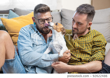 Happy gay couple sitting on floor and playing with dog at home Happy gay couple sitting on floor and playing with dog at home 92622486