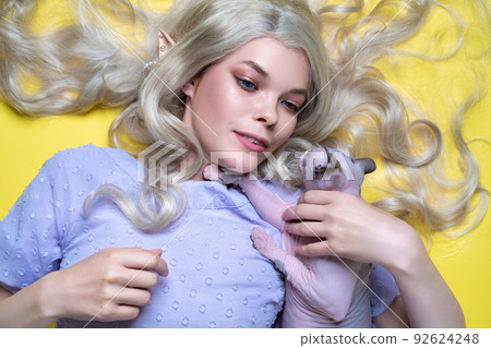 Young blonde woman cosplay elf in blue dress with eyes of different colors lying down on yellow background, holding playful Sphinx kitten on her breasts. Elf has nice curly long hair, pearls in ear. 92624248