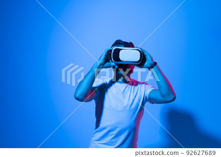 Closeup of young man in white t-shirt wearing virtual reality goggles in modern studio with a neon light. Closeup of young man in white t-shirt wearing virtual reality goggles in modern studio with a neon light. 92627689