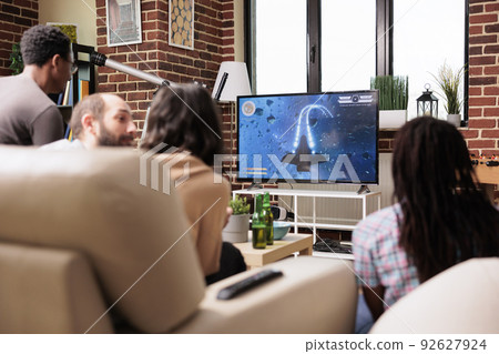 Young adults in living room sitting on sofa while playing space sim videogame on modern console. People sitting at home while enjoying fun leisure activity with electronic entertainment device. Young adults in living room sitting on sofa while playing space sim videogame on modern console. People sitting at home while enjoying fun leisure activity with electronic entertainment device. 92627924