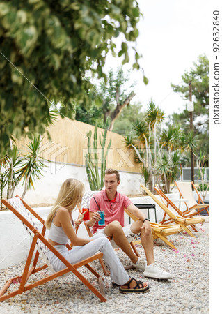 Happy young couple with glasses of lemonade sitting on deck chairs in cozy cafe place with green trees around. Happy young couple with glasses of lemonade sitting on deck chairs in cozy cafe place with green trees around. 92632840