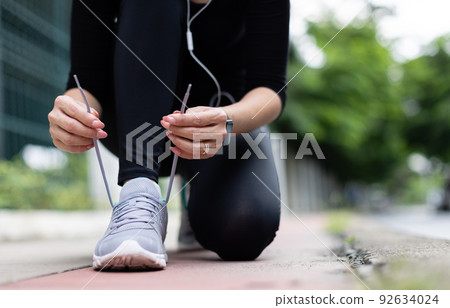 Crop unrecognizable woman runner wearing hygiene protective face mask sitting and tieing shoe rope during workout running in city park. Concept of healthcare in time of coronavirus outbreak 92634024