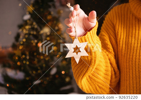 Cropped woman in orange sweater holding Xmas decoration star in front of fir tree with lights on background. Christmas holiday and New Year concept.  92635220