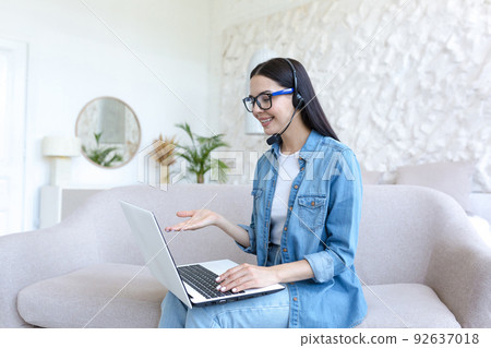 Beautiful woman studying at home, talking on video call with teacher, using headset, woman working sitting on sofa in bright living room 92637018